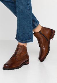 Bottes ankle en cuir marron avec lacets et zip latéral, présentant une texture lisse et une semelle extérieure rugueuse noire avec des accents rouges.