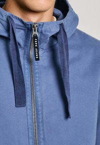 Plava zip-up jakna s kapuljačom napravljena od mekanog materijala, s kontrastnim tamnoplavim vezicama, srebrnim zipom i etiketom s "Pepe Jeans" brandom.