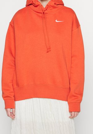 Personne portant un sweat à capuche Nike orange vif avec cordons et logo swoosh blanc, associé à une jupe plissée blanche visible en bas.