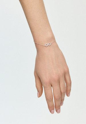 Srebrna bransoletka z łańcuszka z symbolem nieskończoności ozdobionym małymi kamieniami szlachetnymi. Bransoletka noszona jest na dłoni z naturalnymi paznokciami.