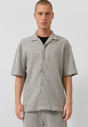 Uomo con capelli corti che indossa un completo abbinato con camicia texturizzata a maniche corte abbottonata e pantaloncini, in piedi davanti a uno sfondo semplice.