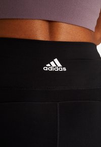 Mustat legginsit korkealla vyötäröllä, joissa on valkoinen Adidas-logo. Materiaali on sileä ja siinä on näkyviä saumat yksityiskohtina.