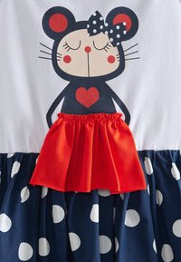 Robe avec un graphique d'un ours avec un cœur sur un haut blanc, accent de volants rouges et une jupe à pois navy. Tissu en mélange de coton.