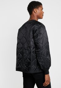 Black quilted jacket with a shiny, textured finish, featuring a round neck and ribbed cuffs, displayed from a side angle.