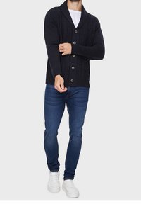 Navy knitted cardigan with a shawl collar, featuring a cable knit pattern and six large buttons. Paired with dark jeans and white sneakers.