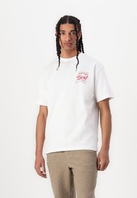 White cotton t-shirt with red "Off the Grid" graphic on the left chest. Crew neck, short sleeves, relaxed fit. Textured tan pants visible.