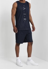 Manlig modell som bär en marinblå ärmlös Nike Air-mesh-top och matchande shorts med vita Nike-sneakers och strumpor mot en vit bakgrund.