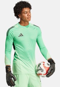 Maglia da portiere verde a maniche lunghe con motivi geometrici, dettagli neri e guanti coordinati, mentre tiene un pallone da calcio con dettagli rossi.