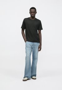 Černé tričko s krátkým rukávem v kombinaci s lehkými modrými zvonovými džíny a bílými teniskami, umístěné na jemném bílém pozadí.