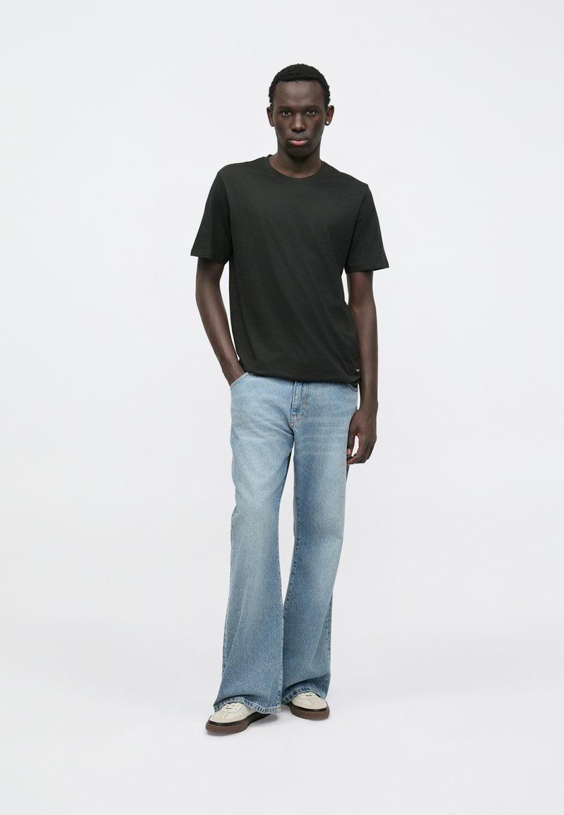 Černé tričko s krátkým rukávem v kombinaci s lehkými modrými zvonovými džíny a bílými teniskami, umístěné na jemném bílém pozadí.