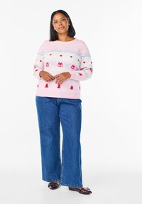 Ružový pletený sveter s červenými a modrými vianočnými vzormi, vrátane srdiečok, darčekov a stromčekov, skombinovaný s modrými širokými džínsami.