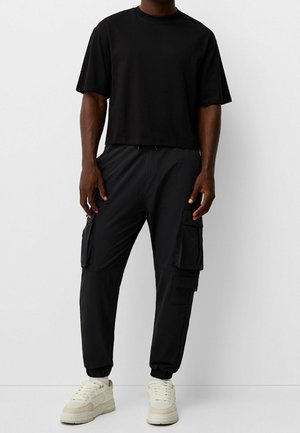 Schwarzes, gekürztes T-Shirt und schwarze Cargo-Hosen mit mehreren Taschen, elastischen Bündchen und einem Kordelzugbund. Weiße Sneakers runden den Look ab.