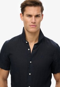 Chłopięca koszula z krótkim rękawem w kolorze granatowym, zapinana na guziki, wykonana z fakturowanej tkaniny, z małym logo na piersi i klasycznym kołnierzykiem.