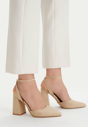 Beżowe zamszowe buty na obcasie typu blok z noskiem w szpic i paskami wokół kostki, noszone z białymi skróconymi spodniami na neutralnym tle.