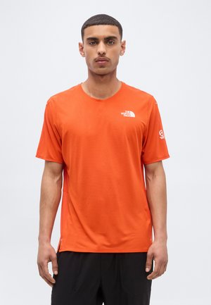 Mann med kortermet knalloransje The North Face T-skjorte og svarte bukser, står mot en ensfarget lys bakgrunn.