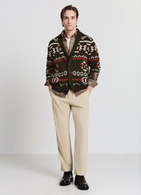 Grün gemusterter Cardigan mit langen Ärmeln und Schalkragen, kombiniert mit einem beigen Hemd und einer beigen Hose, ergänzt durch schwarze Stiefeletten.