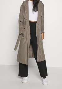 Trench coat lungo in khaki leggero con bottoni, cappuccio regolabile e vita con cintura. Indossato sopra un top bianco corto e pantaloni neri.