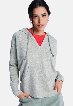 Lichtgrijze capuchontrui met V-hals, trekkoorden en een voorzak, gedragen over een rood shirt. Zachte textuur en een losse pasvorm.