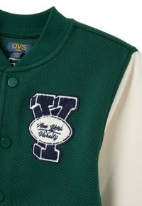 Giacca varsity verde con maniche crema a contrasto, caratterizzata da una superficie testurizzata, chiusura con bottoni e un ricamo "New York Varsity".