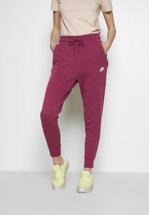 Osoba nosząca bordowe spodnie dresowe Nike ze sznurkiem w pasie i jasnożółte trampki, stojąca na jednolitym białym tle.