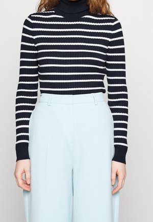 Femme portant un pull à col roulé rayé horizontal noir et blanc et un pantalon large bleu clair taille haute, debout, les bras le long du corps.