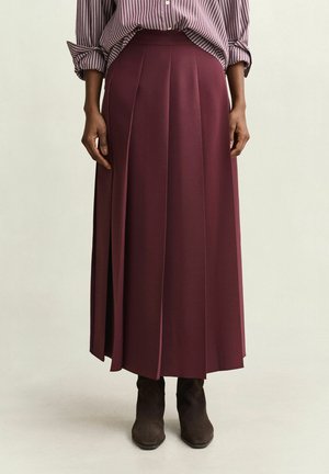 Personne portant une longue jupe plissée bordeaux, une chemise rayée avec les manches retroussées et des bottines foncées sur un fond clair uni.