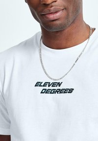Camiseta de algodón blanca con el logo de "ELEVEN DEGREES" en impresión negra y verde azulado; complementada con un collar de cadena plateada.