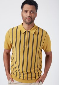 Gul kortärmad polo-tröja med vertikala marinblå ränder, krage och halvdragkedja. Ribbad nederkant och ärmslut. Slät yta.