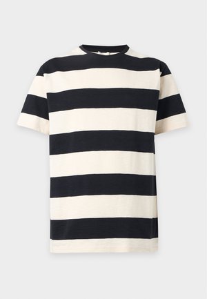Homme debout, les mains dans les poches, portant un t-shirt rayé noir et blanc, un jean large bleu clair et des chaussures marron foncé.