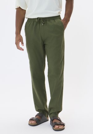 Pantalons en lin vert avec une taille élastique et un cordon de serrage, offrant une coupe décontractée et des poches latérales. Portés avec des sandales sombres.
