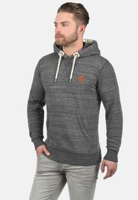 Sweat à capuche gris avec un tissu texturé, poche kangourou, cordons de serrage et petit patch logo marron sur la poitrine. Le sweat à capuche a une doublure intérieure douce.