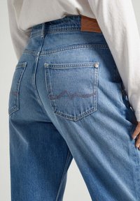 Jeans in denim azzurro con vita alta, dotati di due tasche posteriori, dettagli di cucitura discreti e una toppa in pelle marrone sulla vita.