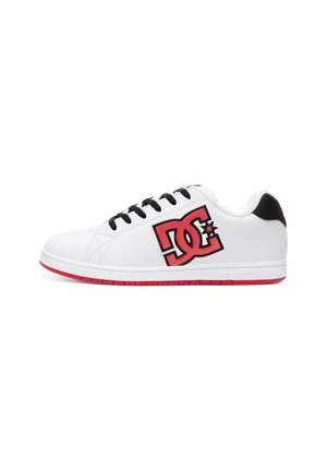 Zapatilla blanca de perfil bajo con cordones negros, suela roja y gran logo DC rojo y negro en el lateral, detalle de cuello de talón negro.