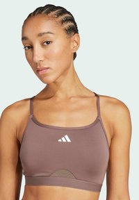 Hnedý športový crop top s tenkými ramienkami, sieťovaným detailom pod prsiami a bielym logom v prednej časti. Hladká elastická tkanina.