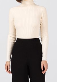 Frau, die einen cremfarbenen Rippenstrickrollkragenpullover trägt, der in hochgeschnittene schwarze weit geschnittene Hosen gesteckt ist, vor einem schlichten Hintergrund.