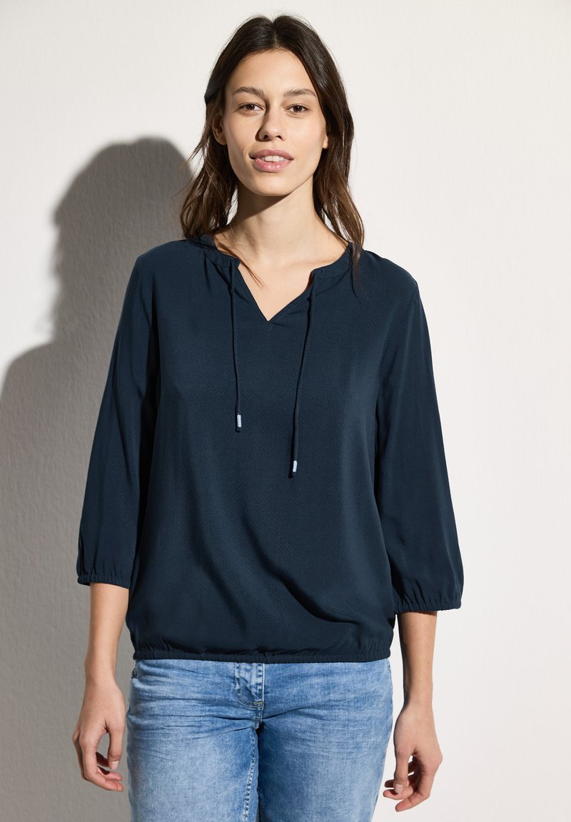 Marineblauwe, lichtgewicht blouse met een V-hals, driekwart mouwen en koorddetail bij de halslijn, gecombineerd met lichtblauwe jeans.