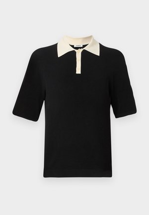 Göndör hajú férfi fekete, rövid ujjú polót, fehér, széllábú nadrágot és bézs cipőt visel, kezében fekete táska van, sima háttér előtt áll.