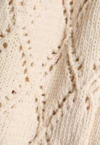 Cremefarbener Strickstoff mit einem strukturierten Muster aus ineinandergreifenden Zöpfen und offenen Maschen, das ein geschichtetes, gewebtes Aussehen erzeugt.