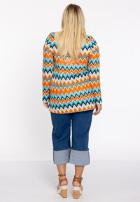 Langermige tuniek met een meerkleurig zigzagpatroon in oranje, turquoise en wit, gecombineerd met denim capri's en sandalen met een plateauzool.