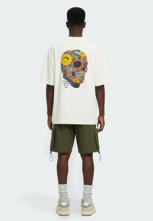 Mies, jolla on valkoinen ylikokoinen T-paita, jossa värikäs pääkallo- ja keltaiset hymynaamakuviot takana, oliivinvihreät cargo-shortsit, valkoiset sukat ja lenkkarit.