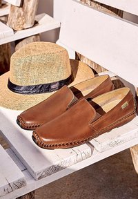 Chaussures en cuir marron à enfiler associées à un chapeau de paille beige avec un ruban noir, posés sur un banc en bois blanc à l'extérieur.