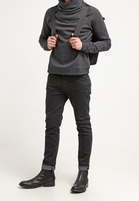 Personne portant un sweat à capuche gris foncé, un jean noir avec revers, des bottes en cuir noires, et portant les bretelles d'un sac à dos noir.