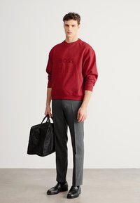 Κόκκινο φούτερ με το λογότυπο «BOSS», γκρι ραμμένο παντελόνι, μαύρα δερμάτινα παπούτσια και μαύρη τσάντα duffel. Ομαλό ύφασμα με άνετη εφαρμογή.