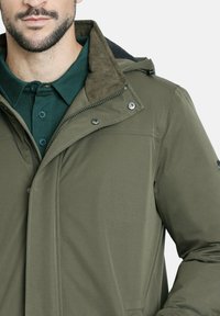 Hombre con una chaqueta verde olivo con cremallera y botones a presión sobre una camisa de color verde oscuro, mostrando el cuello y parte de la cara.