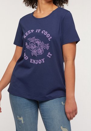Person iført en mørkeblå t-shirt med lilla blomsterdesign og tekst "Keep it cool and enjoy it," kombineret med revnede blå jeans.