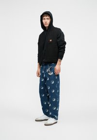 Μαύρο βαμβακερό γιλέκο πάνω από μια μαύρη φούτερ, συνδυασμένο με χαλαρούς μπλε τζιν παντελόνες με σχέδιο γιν-γιάνγκ. Λευκά αθληματικά παπούτσια με γκρίζες λεπτομέρειες.