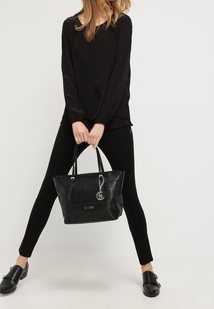 Femme portant un haut, un pantalon et des chaussures noirs, tenant un sac fourre-tout noir Guess avec double poignées et un charme métallique devant elle.