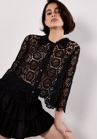Blusa de encaje negro con un patrón floral, cierre de botones y cuello redondo. Tela texturizada y semitranslúcida; mangas largas y corte cropped.