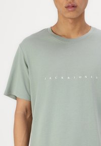 Grønn bomulls T-skjorte med korte ermer og rund hals. Har hvit "JACK & JONES" tekst sentrert på brystet. Myk tekstur.