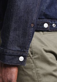 Denim jacket sleeve in dark blue with orange stitching, featuring silver button hardware; worn over olive green fabric, highlighting texture contrast.